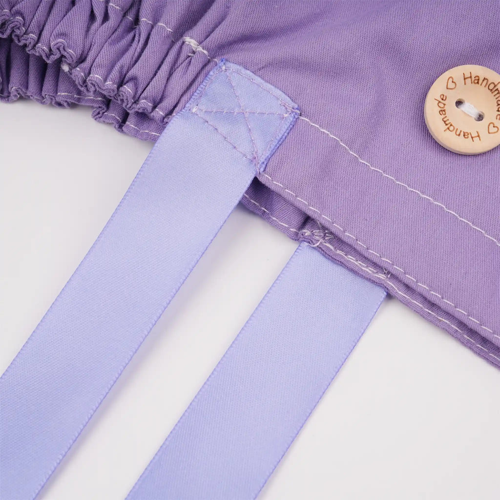 Close-up of light purple scrub cap with a gathered elastic edge, white stitching, a round wooden button engraved with 'Handmade', and two long lavender ribbon ties.