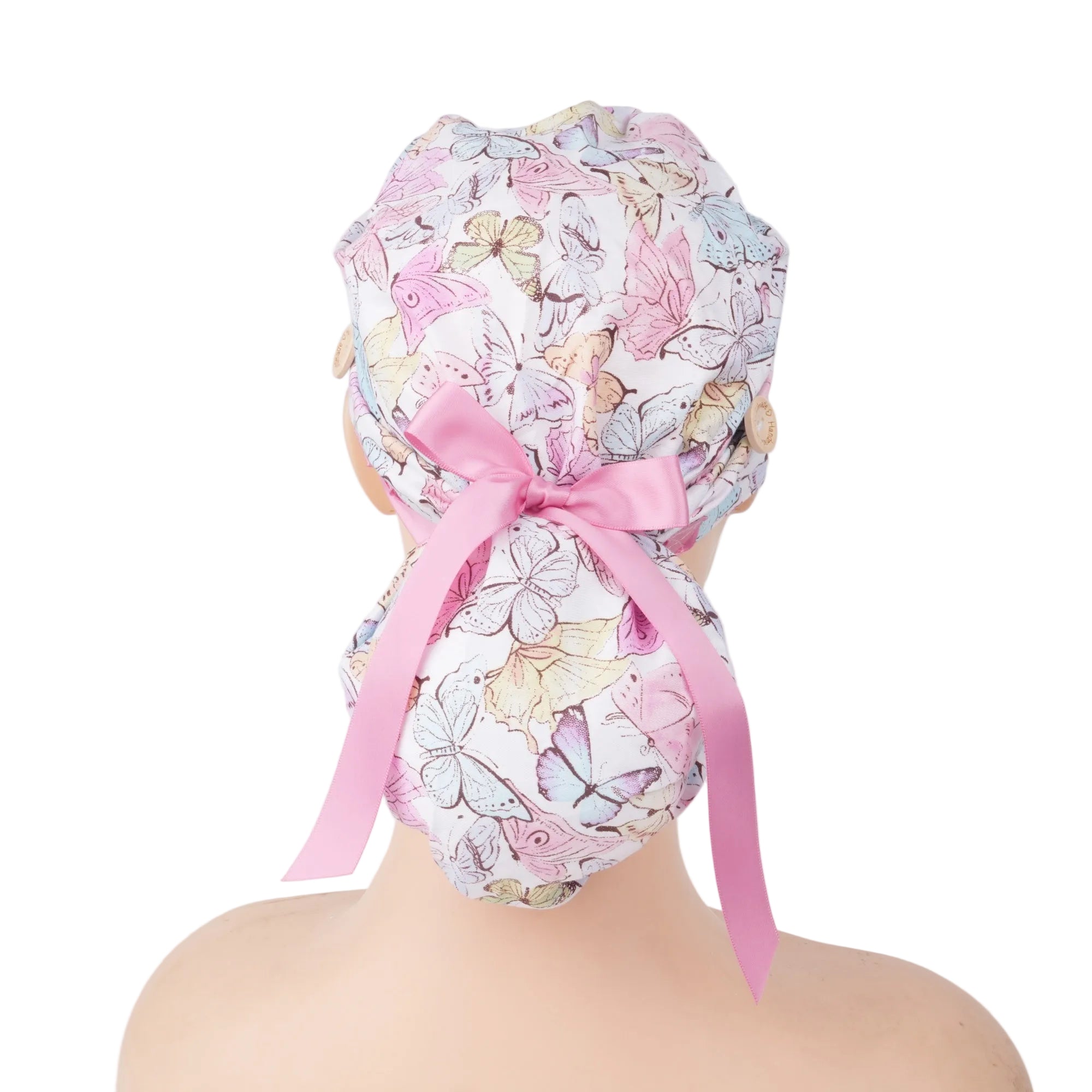 A white fabric scrub cap with a pastel butterfly print and a pink ribbon bow, shown from the back on a mannequin head.