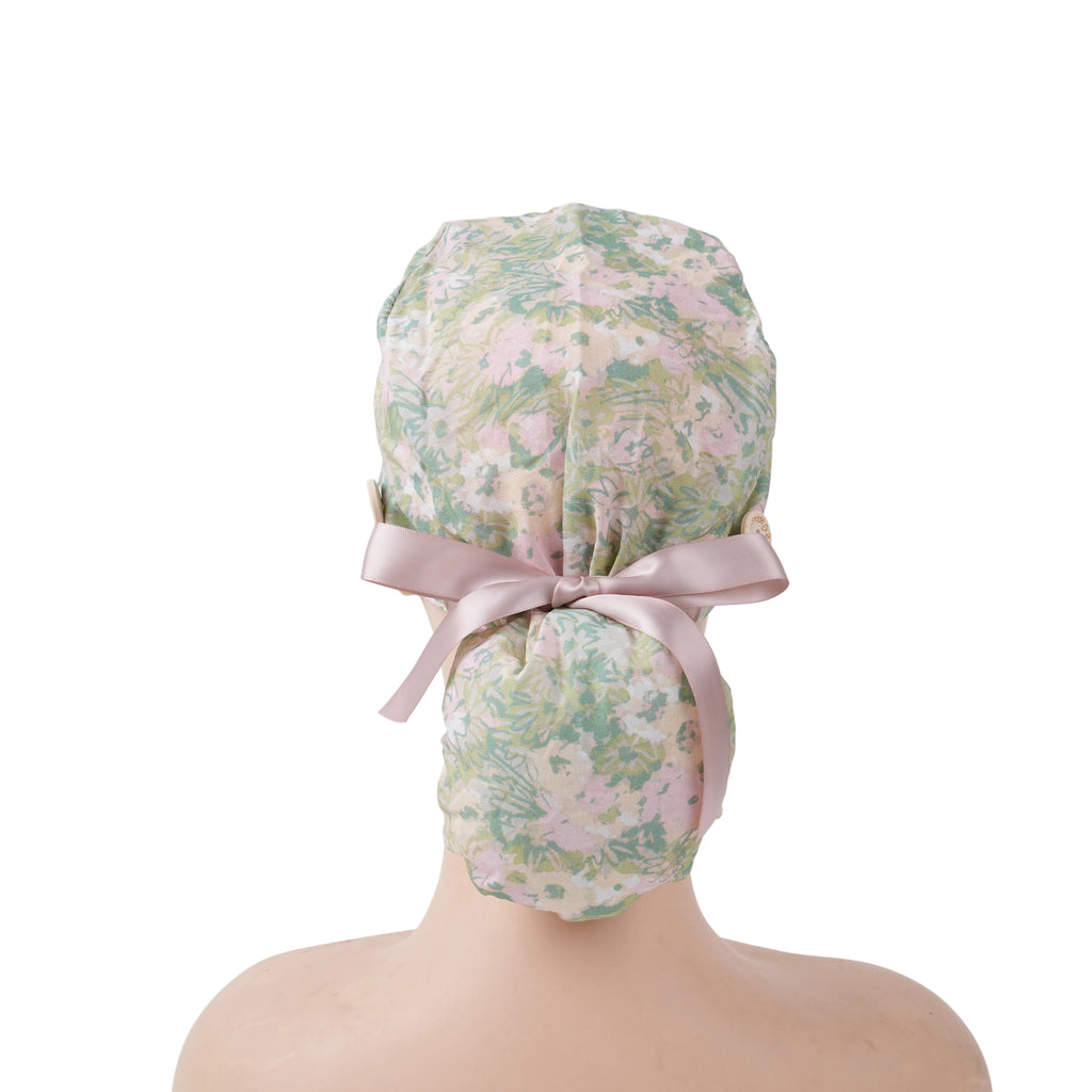 A ponytail scrub cap with floral pattern in green, pink, and beige, with a soft pink satin ribbon at the back, displayed on a mannequin.