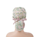 A ponytail scrub cap with floral pattern in green, pink, and beige, with a soft pink satin ribbon at the back, displayed on a mannequin.